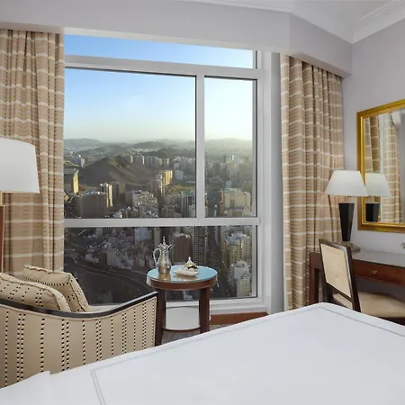 فندق سويس اوتيل المقام مكة المظهر الخارجي الصورة The photo shows a well-furnished hotel room featuring a large window with a view of a cityscape and mountains in the background. On the left, there is a comfortable armchair next to a side table with two glasses and a bottle. In front of the window, 